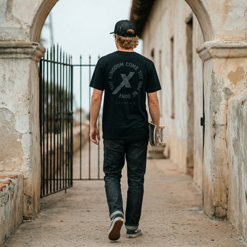 Man wearing Black Thy Kingdom Come Catholic Shirt, back print, walking through a church arch